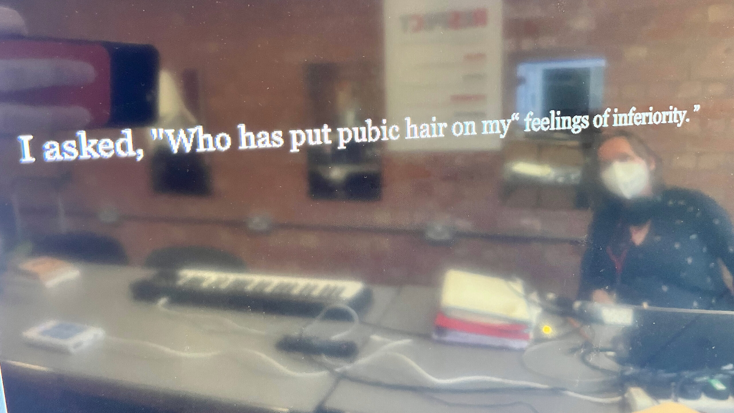 A photo of the composer Sarah Angliss reflected in a TV screen, which displays the text "I asked, "Who has put public hair on my feelings of inferiority.